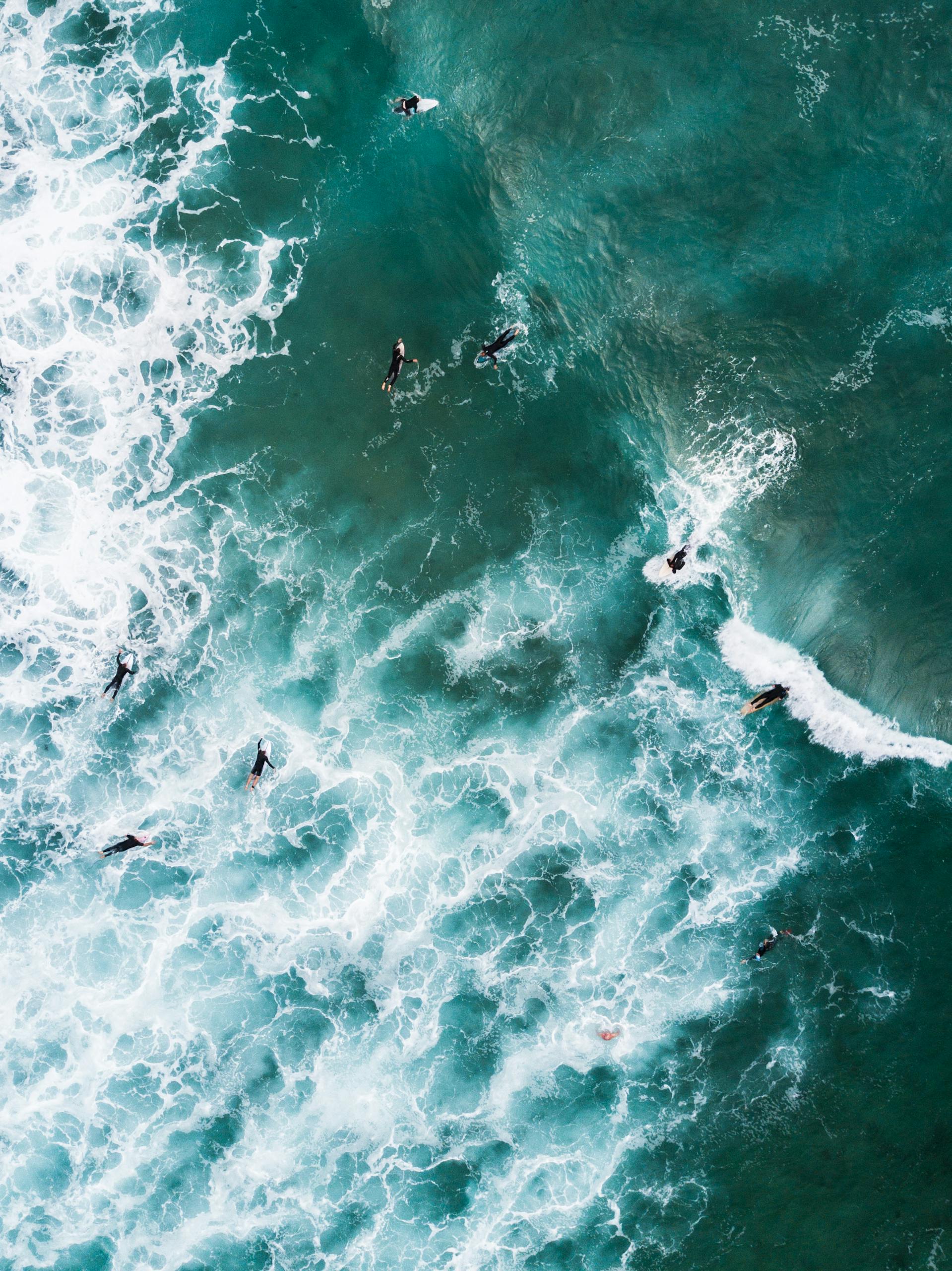 Aerial shot of surfers catching ocean waves with vibrant seafoam and clear water.
